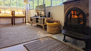 A cozy room with a large window, a sofa with yellow cushions, a wooden entertainment unit, a woven basket, and a wood-burning stove. - Cottage | Pool | Mountain View | Jacuzzi (Arganil)