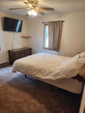 Un dormitorio con una cama, ventilador de techo, televisor montado en la pared y ventana con cortinas.
