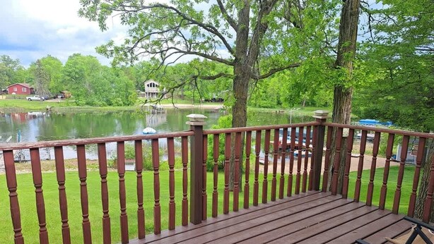 Una terraza de madera con barandilla que da a un lago, rodeada de vegetación y una casa al fondo.