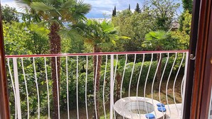 Ein Balkon mit weißer Brüstung, ein runder Tisch mit blauen Kissen und üppiges Grün im Hintergrund.