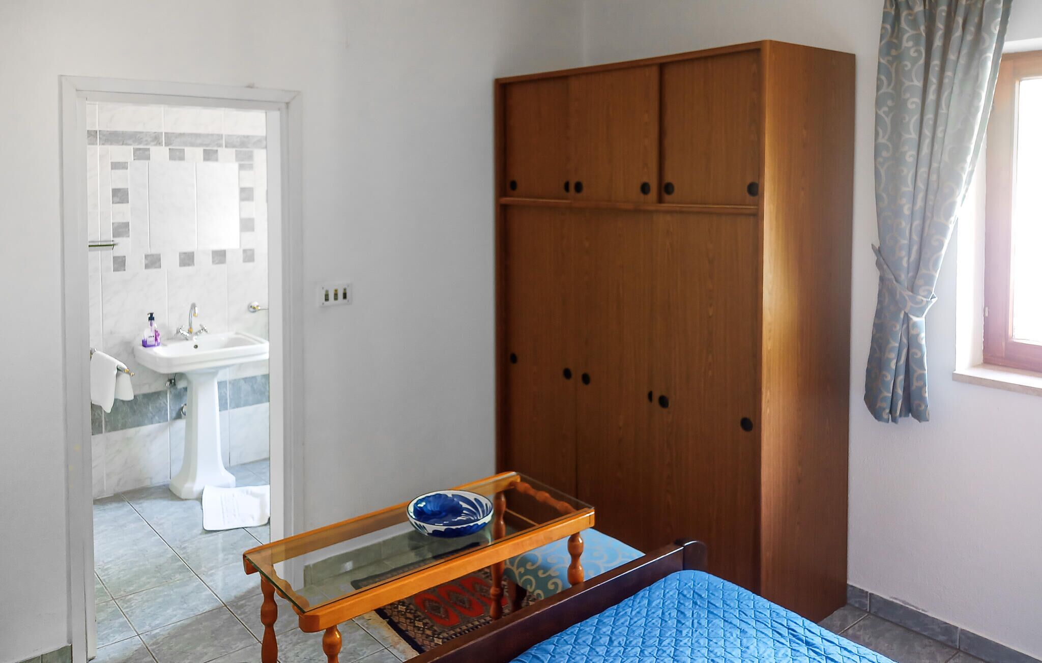 Un dormitorio con cama, un armario de madera, una mesita de cristal con un cuenco, un baño con lavabo y una ventana con cortinas.