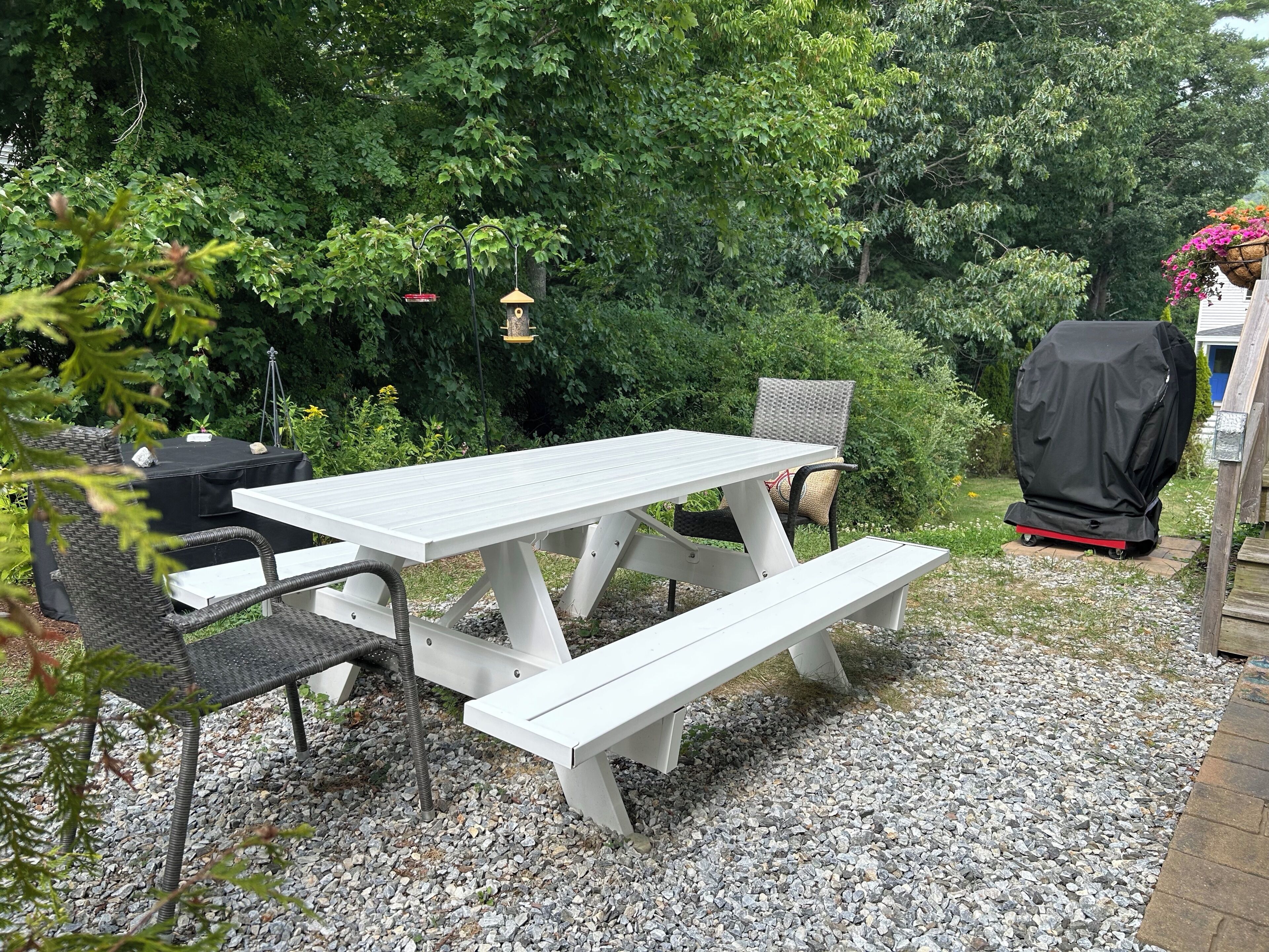 Une table de pique-nique blanche avec des bancs, entourée de gravier, avec un barbecue et des mangeoires à oiseaux en arrière-plan.