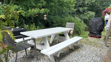 Une table de pique-nique blanche avec des bancs, entourée de gravier, avec un barbecue et des mangeoires à oiseaux en arrière-plan.