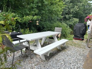 Ett vitt picknickbord med bänkar, omgivet av grus, med en grill och fågelmatare i bakgrunden.