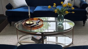 Un sofá azul con almohadones a rayas, una mesa de centro de cristal con un jarrón de flores amarillas, y vistas a un jardín verde a través de amplios ventanales.