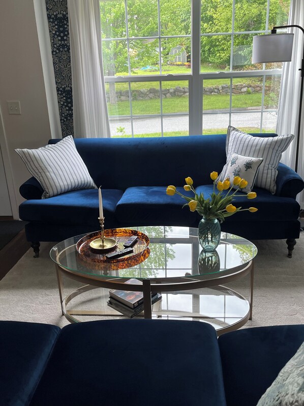 Un sofá azul con almohadones a rayas, una mesa de centro de cristal con un jarrón de flores amarillas, y vistas a un jardín verde a través de amplios ventanales.
