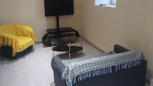 A room with a sofa covered in yellow, a blue sofa with a checkered blanket, a TV on a stand, and a window. - Charming country house (Cours)