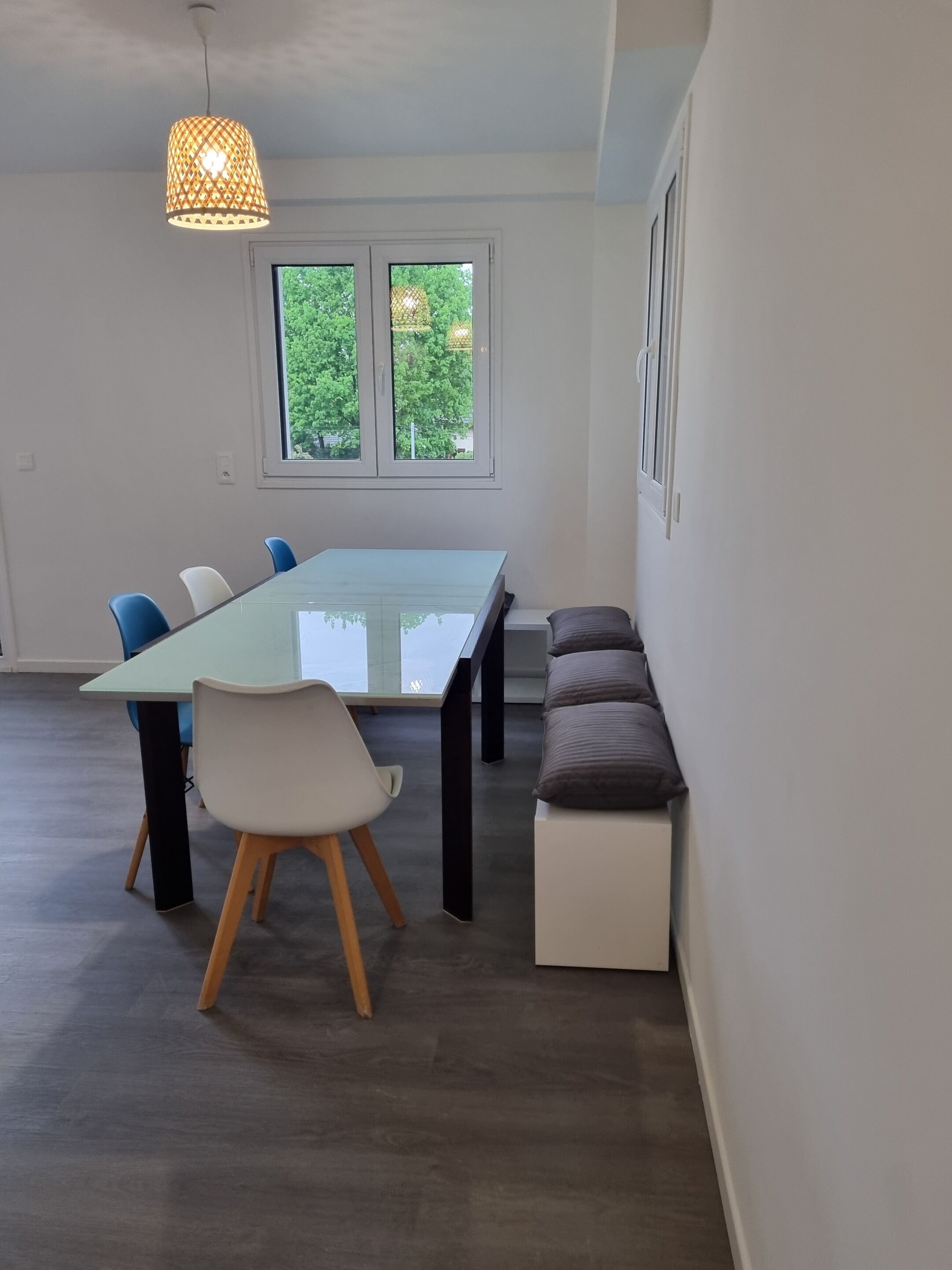 A modern room with a glass-top dining table, blue and white chairs, a bench with cushions, a hanging light fixture, and a window with a view of greenery.