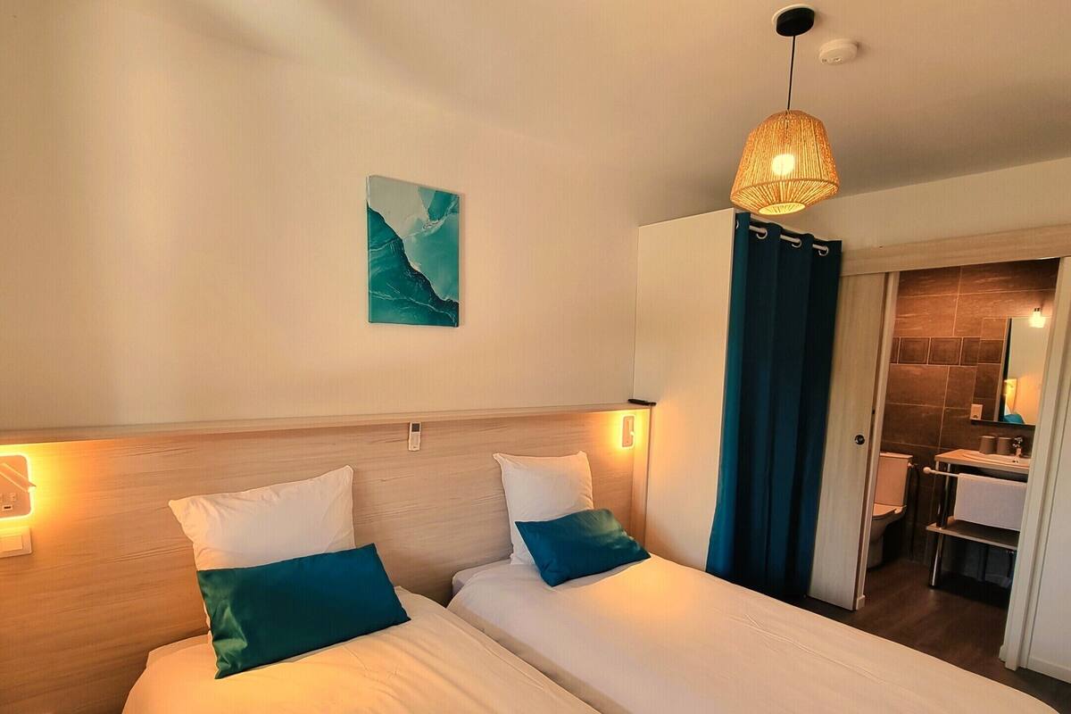 Ein Hotelzimmer mit zwei Betten, einem Bild an der Wand, einer Pendelleuchte und einem durch eine offene Tür einsehbaren Badezimmer.