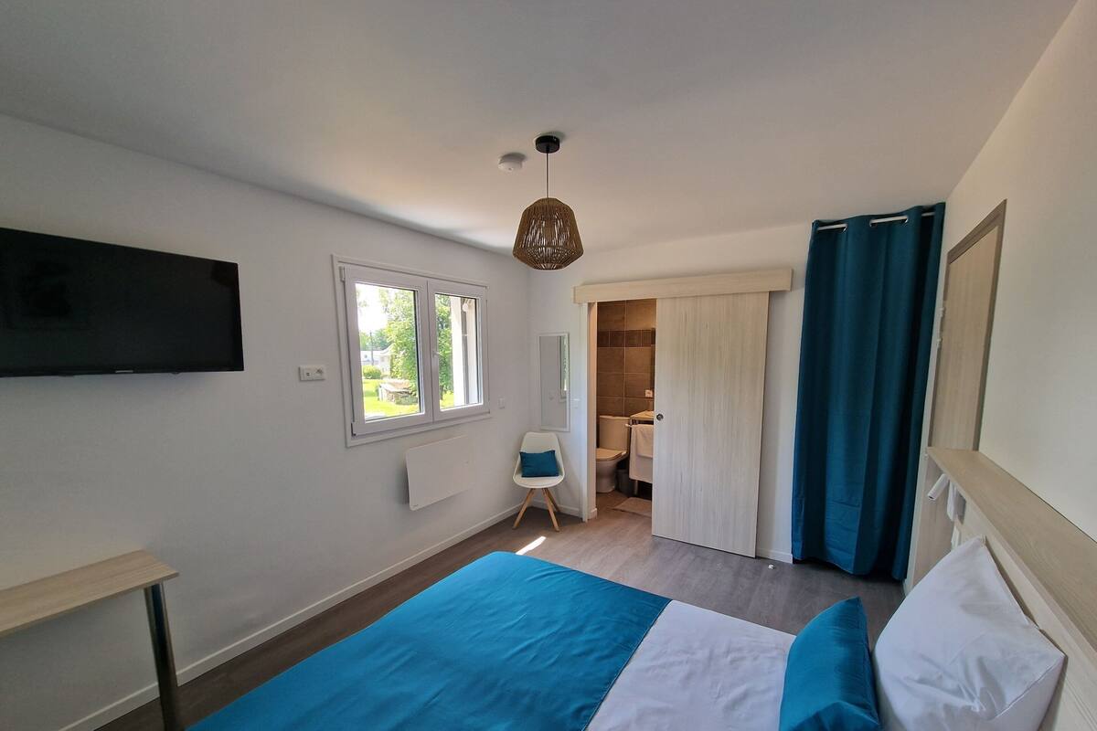 Ein Schlafzimmer mit Bett, einem an der Wand montierten Fernseher, einem Fenster und einem durch eine offene Tür einsehbaren Badezimmer.