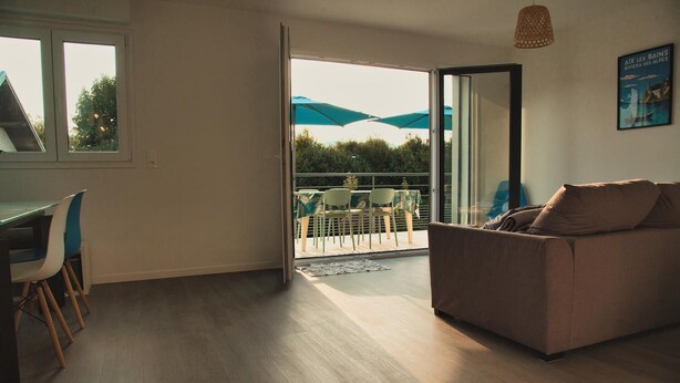 An open-plan living area with a sofa, dining table, and chairs. A sliding glass door leads to an outdoor terrace with umbrellas. - Le Combo (Aix-les-Bains)