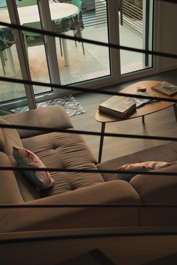 A modern living room with a sofa, a coffee table, and a view through large windows to an outdoor area with a table and chairs. - Le Combo (Aix-les-Bains)