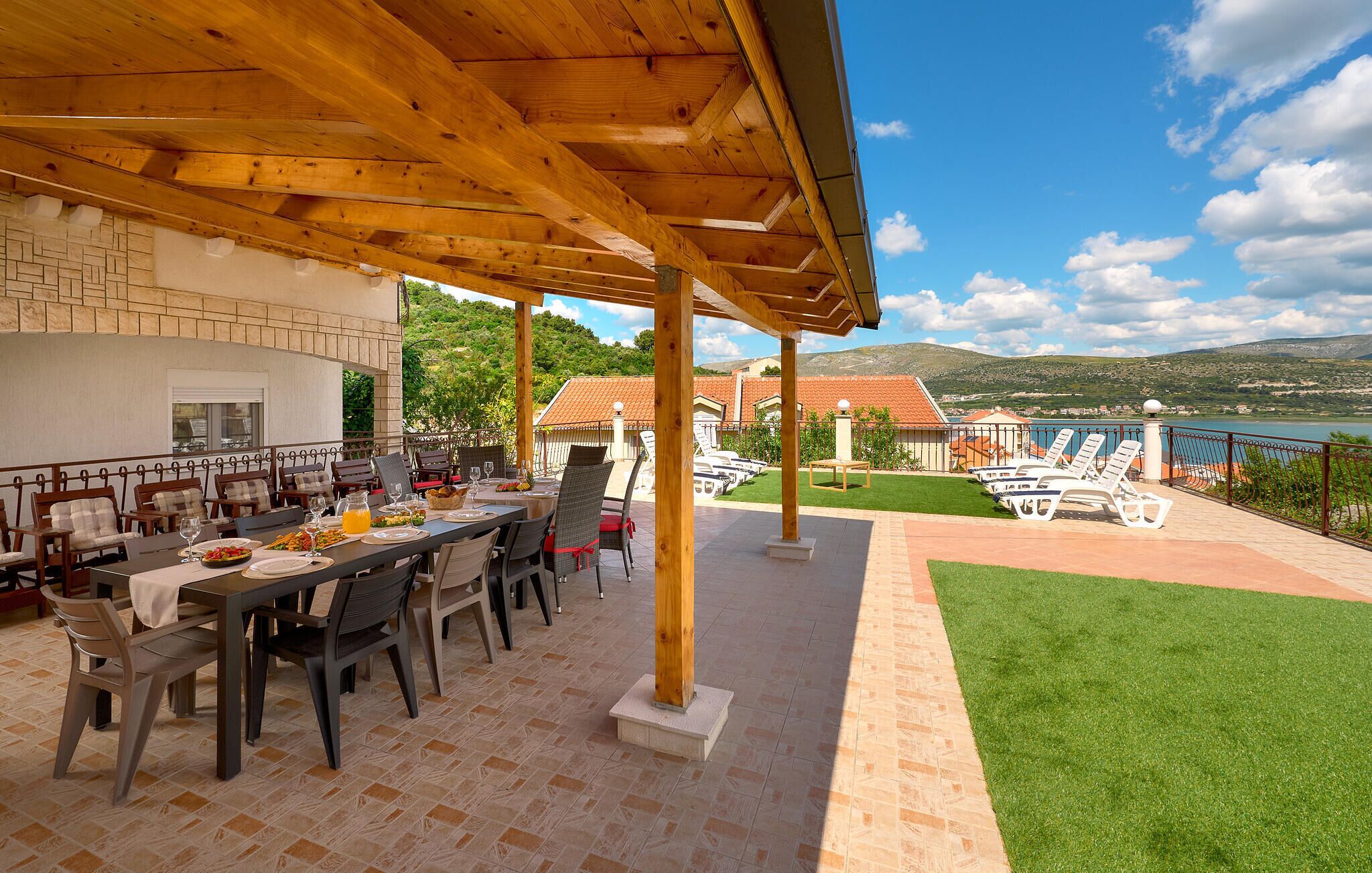 An outdoor dining area with a wooden pergola, a table set for a meal, and a view of a lake and hills in the background.
