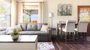 A cozy living room with a grey sofa, a coffee table with a plant, a dining area with chairs, and a large window with curtains. - Bluejay Bungalow – Cozy Pet-Friendly Cottage Near Yosemite & Bass Lake (Oakhurst)