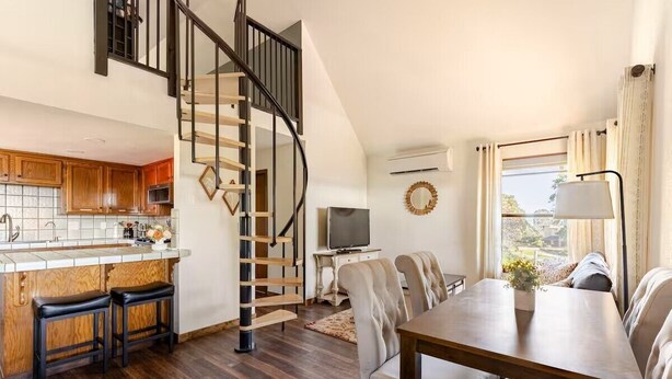 A modern kitchen with wooden cabinets, a built-in island with stools, a spiral staircase, a dining area with a table and chairs, and a TV in the living room. - Bluejay Bungalow – Cozy Pet-Friendly Cottage Near Yosemite & Bass Lake (Oakhurst)