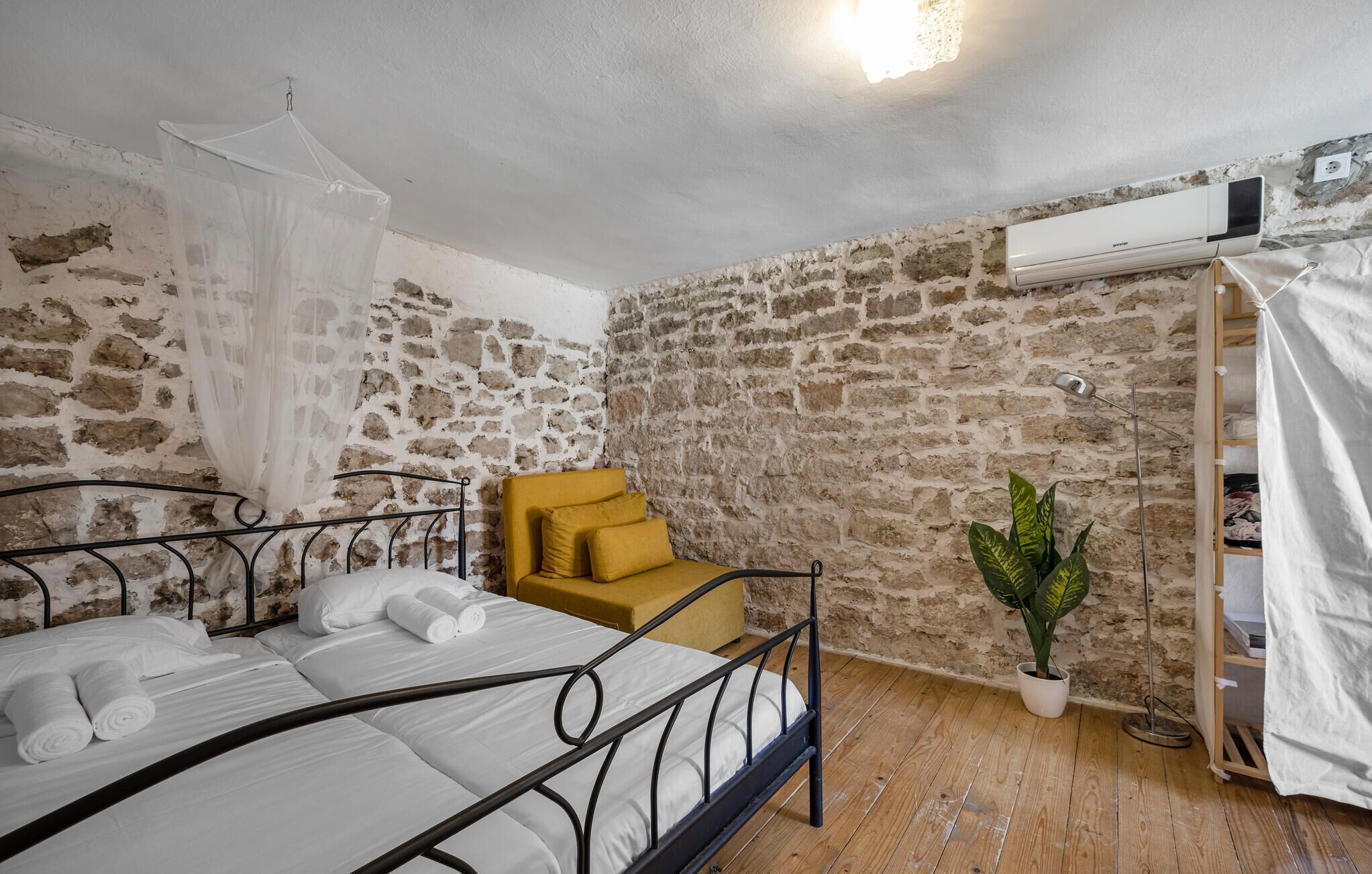 Una camera da letto con un letto, una poltrona gialla, una pianta in vaso e un muro in pietra.