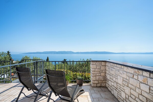 A balcony with two chairs, a potted plant, and a stone wall overlooking a body of water and distant mountains.