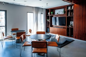 Ein modernes Interieur mit einem runden Tisch, orangenen Stühlen, einem Holzregal und einem großen Fenster.