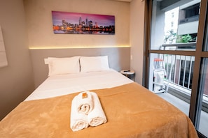 A hotel room with a bed, a nightstand, a painting of a cityscape, and a window with a view of buildings.