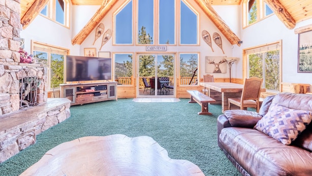 A cabin interior with a stone fireplace, a large TV, wooden furniture, and a view of the outdoors.
