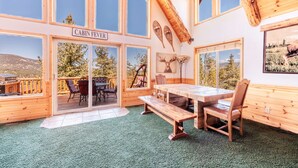 A cabin interior with a large wooden table, benches, and a view of the outdoors through large windows.