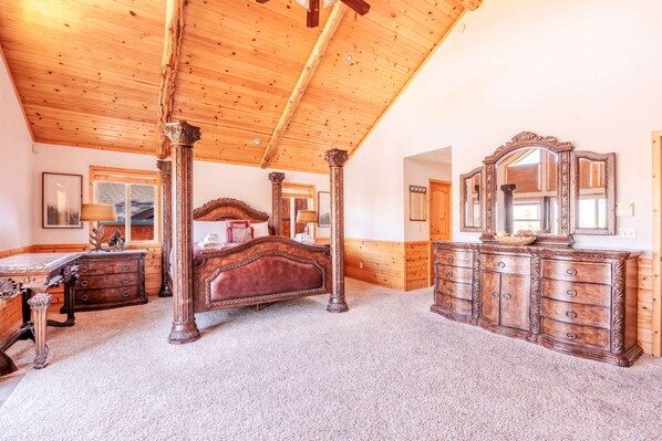 A bedroom with a wooden ceiling, a four-poster bed, a dresser with a mirror, and a carpeted floor.