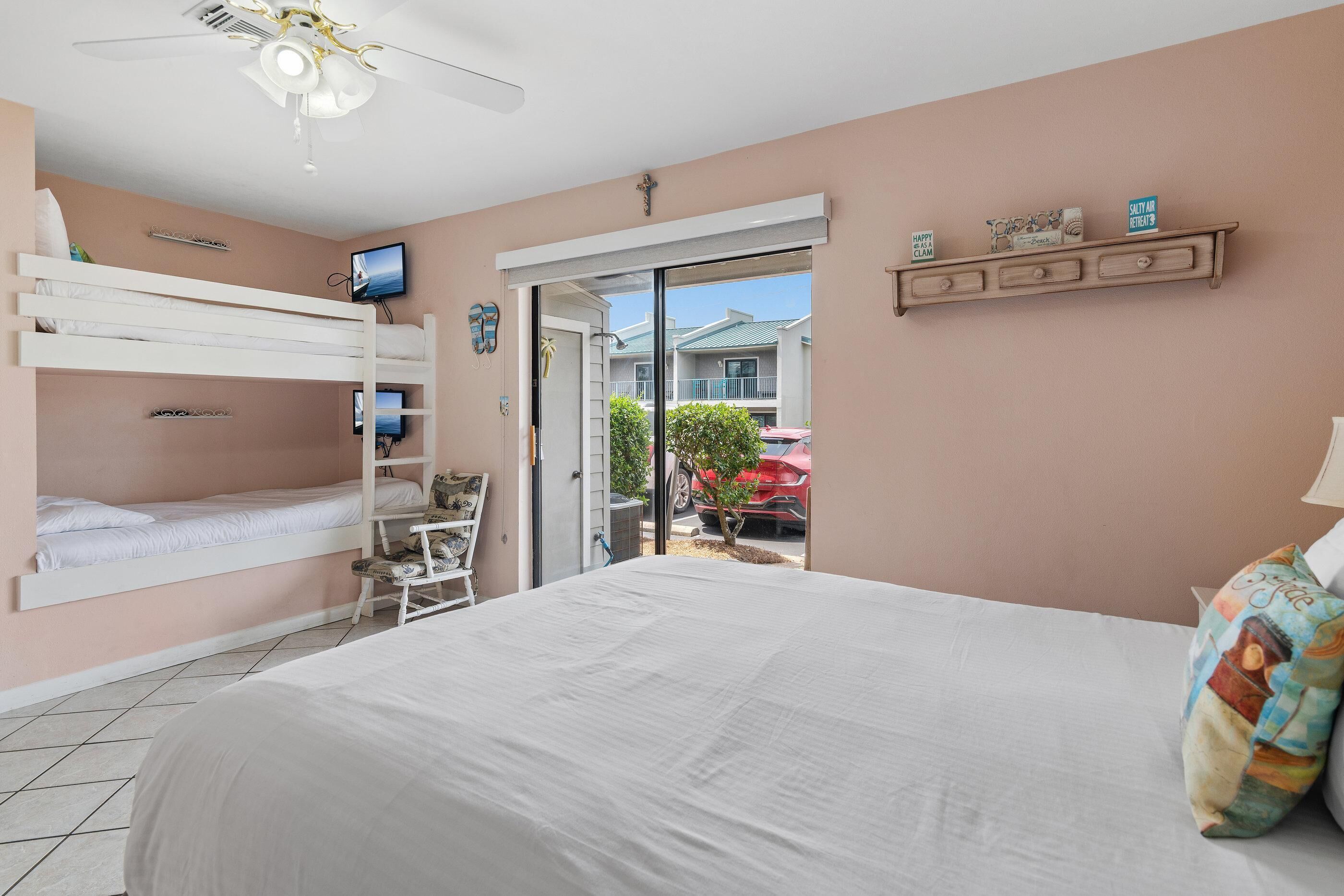 A bedroom with bunk beds, a ceiling fan, a television, and a sliding glass door leading to a balcony with a view of houses and a red car.