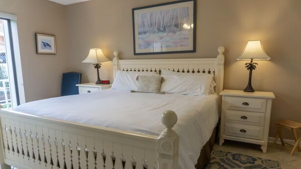 A bedroom with a white bed, two bedside tables with lamps, a small chair, and a framed picture on the wall. - Gulf Winds East #10- The Turtles Nest (Miramar Beach)