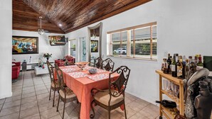 Un comedor con techo de madera, una gran mesa con mantel floral y sillas de respaldo ornamentado. Hay cuadros en las paredes, una ventana con persianas y una estantería con botellas de vino.