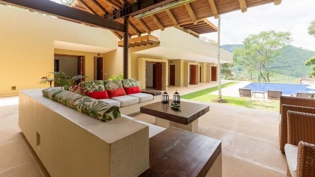 A spacious outdoor seating area with a sofa, coffee table, and wicker chairs, overlooking a pool and lush greenery. - Yeguas W27 by Wynwood House (Anapoima)