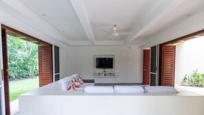 A modern living room with a white sofa, a flat-screen TV mounted on the wall, and sliding wooden doors leading to an outdoor area. - Yeguas W27 by Wynwood House (Anapoima)