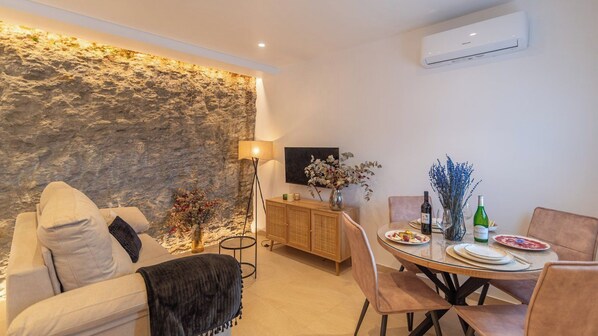 A modern living room with a stone wall, a sofa, a dining table set with plates and wine bottles, and a flat-screen TV.