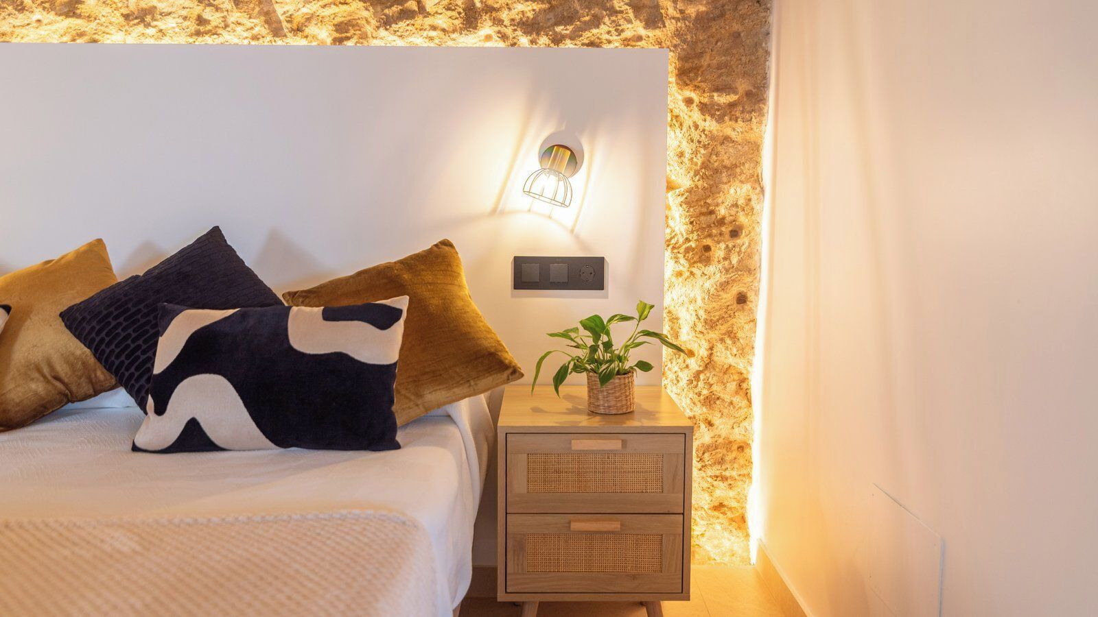 A neatly made bed with a white comforter and several pillows, a bedside table with a plant, and a wall-mounted light fixture.
