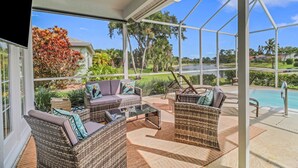 Eine überdachte Terrasse im Freien mit Korbmöbeln, einem Glastisch und Blick auf einen Swimmingpool und die Landschaft.