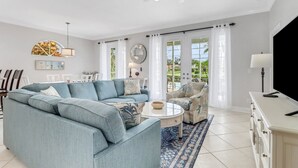 Ein geräumiges Wohnzimmer mit einem hellblauen Ecksofa, einem runden Couchtisch und einem gemusterten Sessel. Es gibt einen Essbereich mit einem weißen Tisch und Stühlen, und ein großes Fenster mit Blick auf Grünflächen außerhalb.