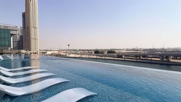Une piscine à débordement située sur le toit, avec une eau d’un bleu cristallin, des chaises longues blanches et une vue sur un grand immeuble et un pont en arrière-plan.