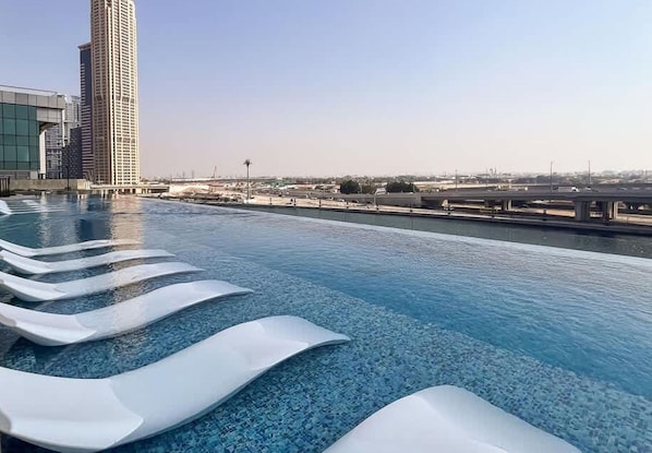 A rooftop infinity pool with lounge chairs, a view of a tall building, and a bridge in the background.