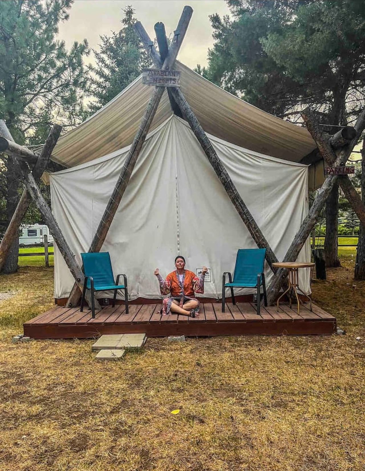 A tent with a wooden frame and white fabric walls, a wooden deck, two blue chairs, and a person sitting on the deck.