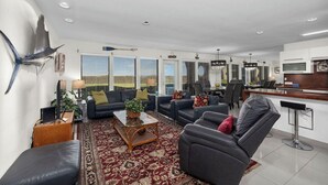 Una sala moderna con sofá, sillón y mesa de centro, un área de cocina con mostrador y electrodomésticos, y ventanas amplias con persianas.