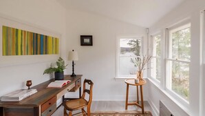 Quarto com escrivaninha de madeira, cadeira, planta em vaso, luminária e janela com vista para árvores.