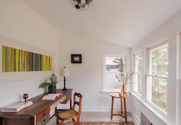 Een kamer met een houten bureau, een stoel, een potplant, een lamp en een raam met uitzicht op bomen.