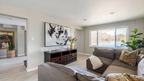 Uma sala de estar espaçosa com uma grande janela, um sofá moderno, uma mesa de centro e um objeto decorativo na parede.