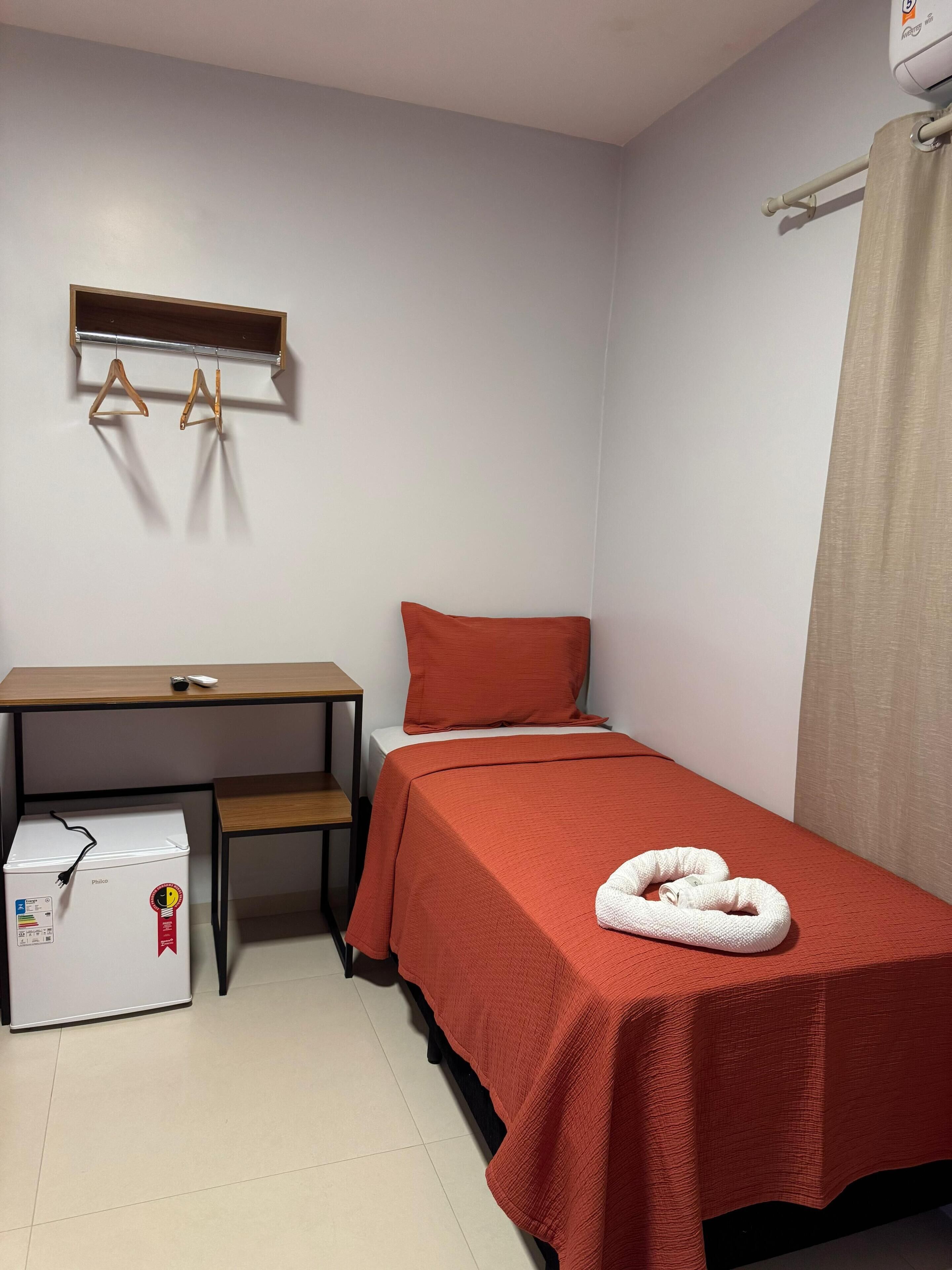 A small, neatly arranged room with a bed, a desk, a chair, a heater, and a curtain.