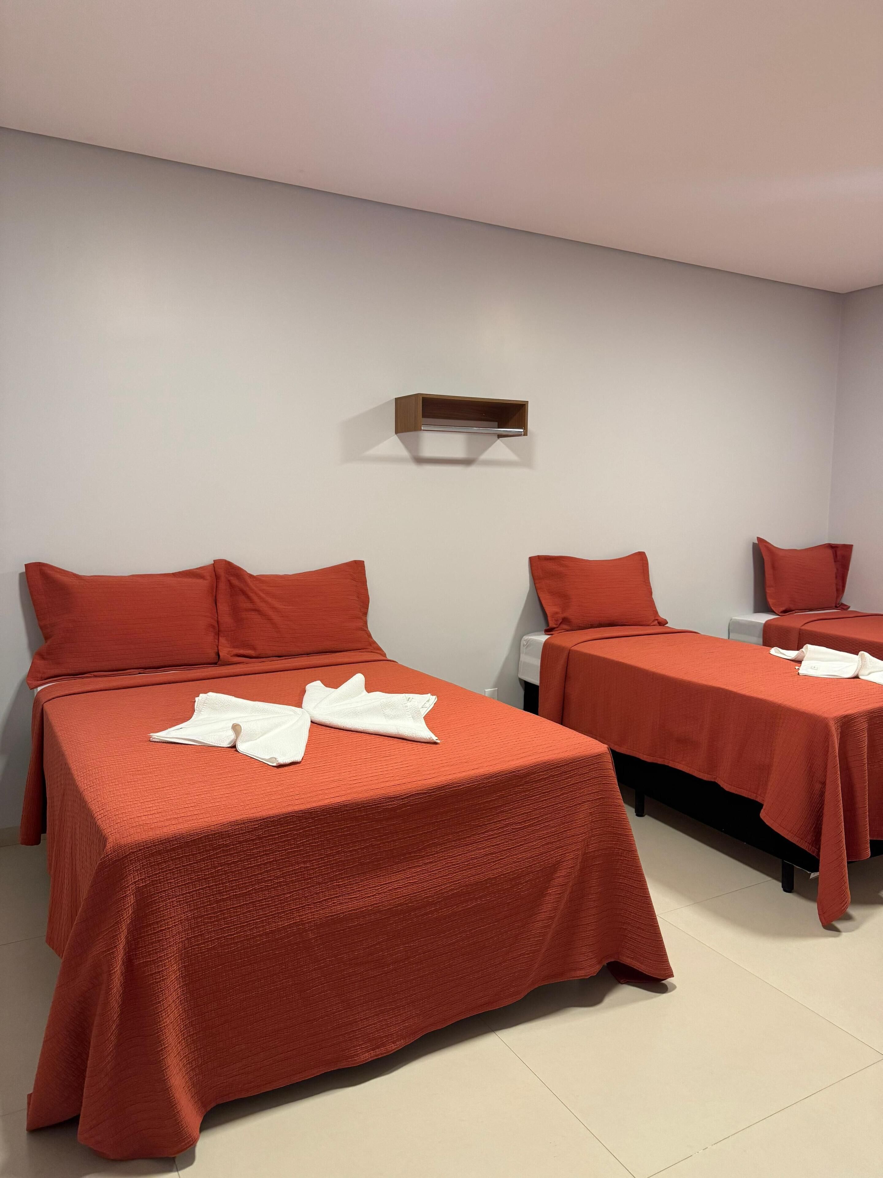A hotel room with two beds, each with a red bedspread and matching pillows. There is a small wooden shelf above the beds.