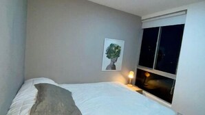 A bedroom with a bed, a nightstand, a wall-mounted lamp, and a window with a view of the night sky.