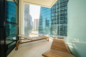 A balcony with a wooden lounge chair and a small table, overlooking a cityscape with modern buildings.