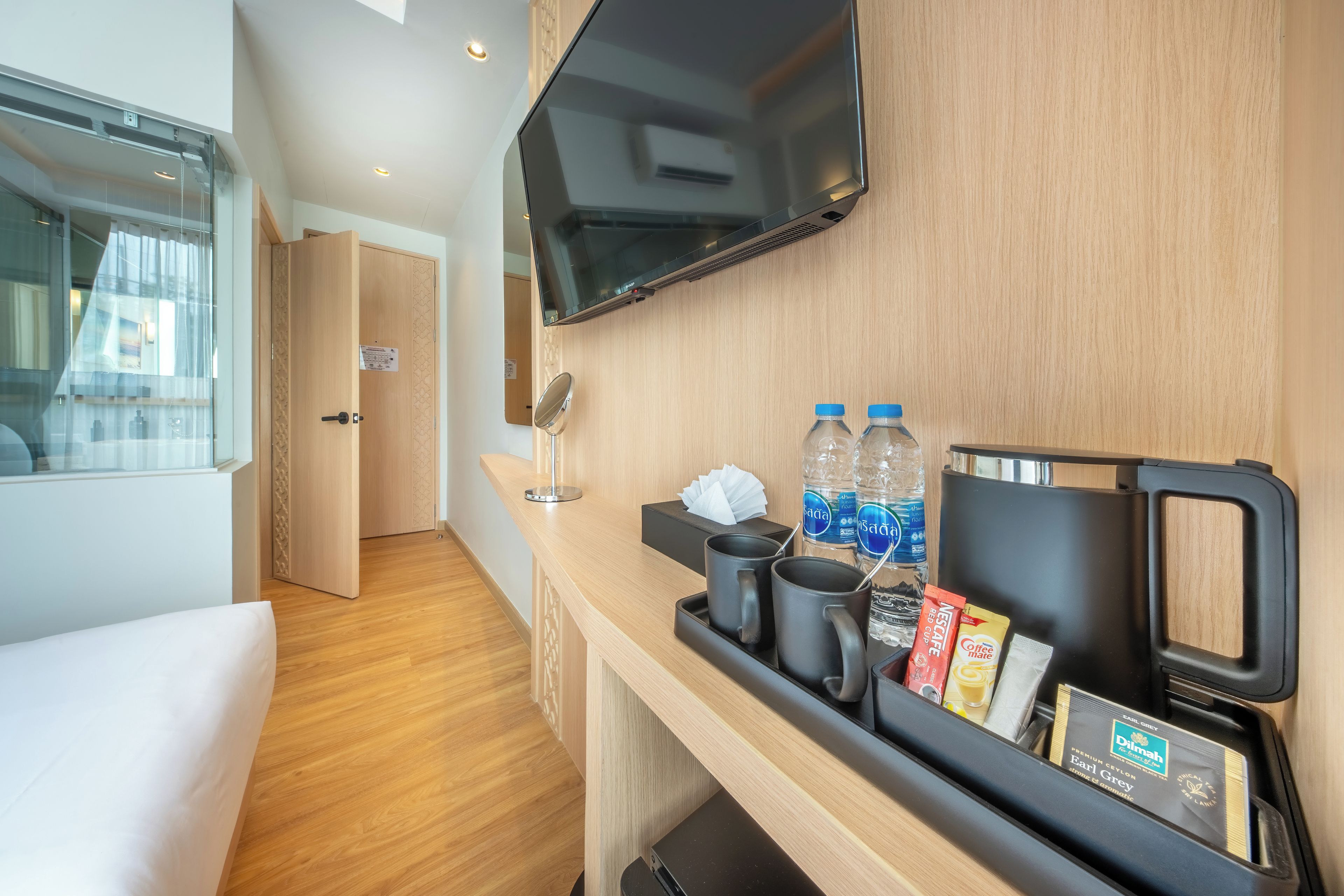 Habitación de hotel con piso de madera, una cama, un televisor montado en la pared, una mesita con agua embotellada, café y té, y un espejo pequeño.