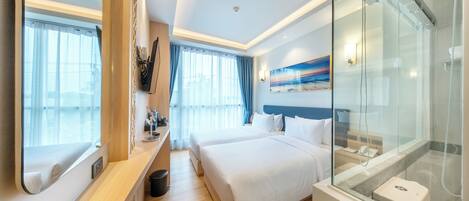 Une chambre d’hôtel avec un grand lit, une salle de bain séparée dotée d’une douche en verre, et une vue sur la mer depuis la fenêtre.
