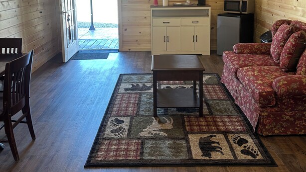 A cozy cabin interior with a wooden ceiling, a ceiling fan, a dining area with a table and chairs, a sofa, a kitchen area with cabinets and appliances, and a large window. - Cabin 1 in peaceful Reed Point 3 minutes from Yellowstone River (Reed Point)