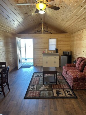 A cozy cabin interior with a wooden ceiling, a ceiling fan, a dining area with a table and chairs, a sofa, a kitchen area with cabinets and appliances, and a large window. - Cabin 1 in peaceful Reed Point 3 minutes from Yellowstone River (Reed Point)